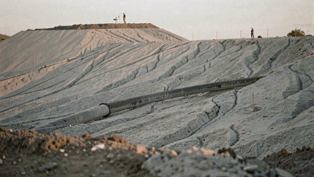 A dewatered tailings stack seen from a low ridge at late afternoon, the surface a pale grey crust with shallow erosion channels, a single discharge pipe crossing the foreground and a line of survey