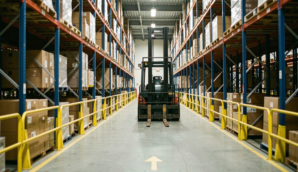 A high-bay pallet racking aisle at floor level, one reach truck parked with forks lowered, yellow guard rail separating a marked pedestrian walkway, cold overhead lighting from roof-mounted LED